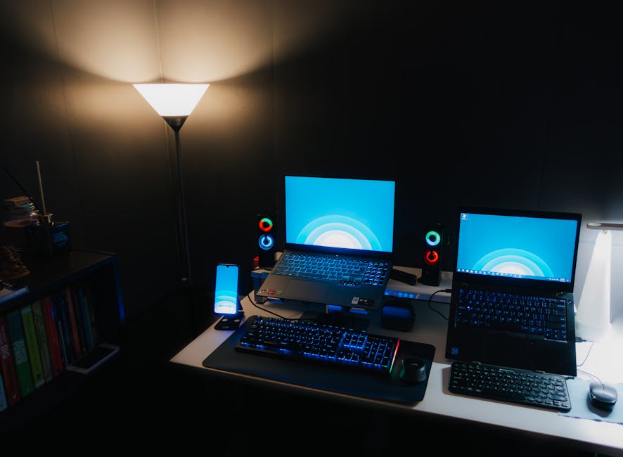 Nighttime office desk setup with laptops, illuminated keyboard, and ambient lighting.